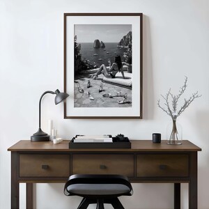 May include: A framed black and white wall art print depicting a coastal scene with a person in a bikini lounging on a terrace with scattered high-heeled shoes, displayed above a dark wooden desk with a black lamp and a glass vase with branches.