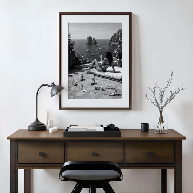 May include: A framed black and white wall art print depicting a coastal scene with a person in a bikini lounging on a terrace with scattered high-heeled shoes, displayed above a dark wooden desk with a black lamp and a glass vase with branches.