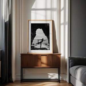 K&ouml;nnte beinhalten: Schwarz-Wei&szlig;-Druck mit Rahmen einer Person, die eine Yoga-Pose vor dem Meer ausf&uuml;hrt. Das Bild ist in einem hellen Holzrahmen gerahmt und wird auf einem dunklen Holzschrank ausgestellt. Der Hintergrund umfasst ein Fenster und eine graue Wand.