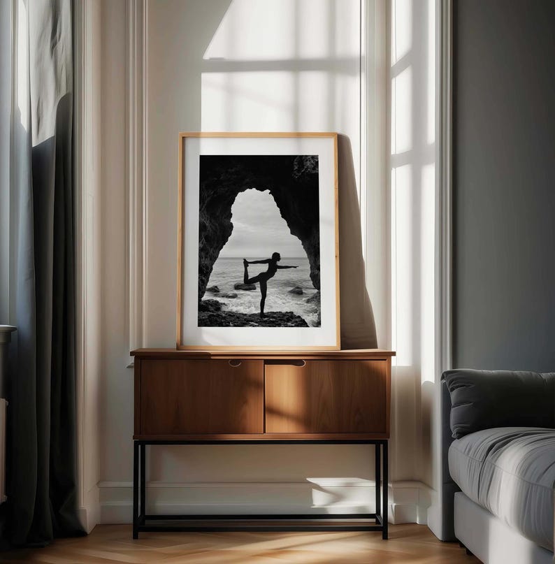 K&ouml;nnte beinhalten: Schwarz-Wei&szlig;-Druck mit Rahmen einer Person, die eine Yoga-Pose vor dem Meer ausf&uuml;hrt. Das Bild ist in einem hellen Holzrahmen gerahmt und wird auf einem dunklen Holzschrank ausgestellt. Der Hintergrund umfasst ein Fenster und eine graue Wand.