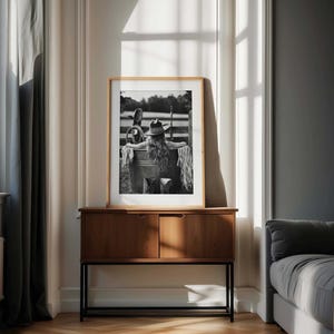 May include: Monochrome framed print of a person in a cowboy hat and fringe, leaning against a metal tub. The print is displayed on a wooden cabinet with a black metal base, in a room with a window.