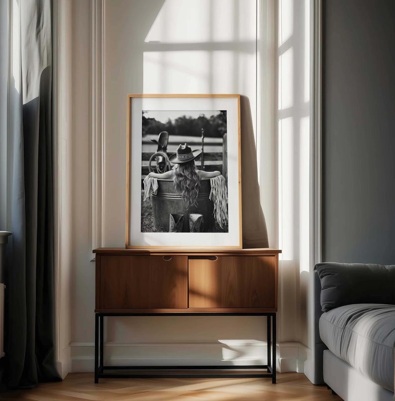 May include: Monochrome framed print of a person in a cowboy hat and fringe, leaning against a metal tub. The print is displayed on a wooden cabinet with a black metal base, in a room with a window.