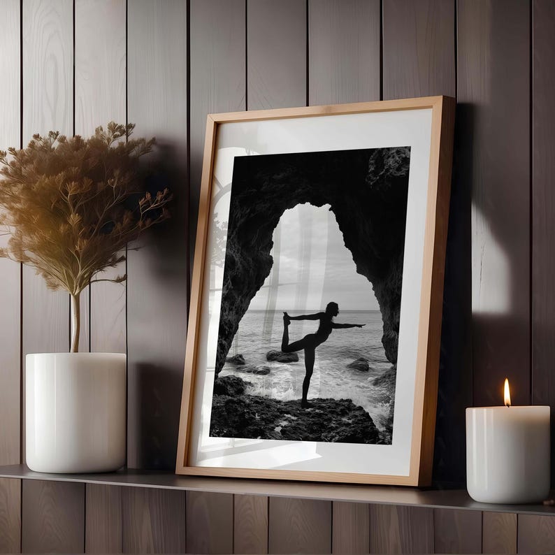 K&ouml;nnte beinhalten: Schwarz-Wei&szlig;-Druck in einem Rahmen, der eine Person beim Yoga an einem felsigen Ufer zeigt, eingerahmt von einem nat&uuml;rlichen Torbogen. Das Bild vor dem Hintergrund des Meeres und des Himmels. Eine wei&szlig;e Kerze und eine Topfpflanze stehen auf einem Regal.