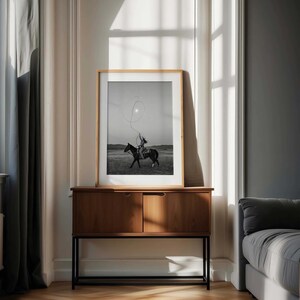 May include: A framed black and white photograph depicting a person riding a horse while swinging a lasso in an open field. The artwork is displayed in a light-colored wooden frame, resting on a mid-century modern wooden cabinet with black legs.