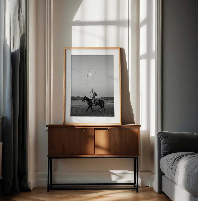 May include: A framed black and white photograph depicting a person riding a horse while swinging a lasso in an open field. The artwork is displayed in a light-colored wooden frame, resting on a mid-century modern wooden cabinet with black legs.