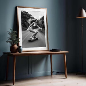 Peut inclure: Tirage encadr&eacute; en noir et blanc d'une personne faisant du skateboard sur une route. La personne porte un maillot de bain et a de longs cheveux. L'impression est dans un cadre en bois, sur une table en bois.