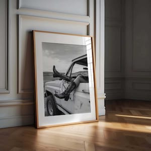 Pu&ograve; includere: Fotografia in bianco e nero incorniciata di stivali da cowboy che sporgono dal finestrino di un camion bianco. Gli stivali hanno cuciture decorative. Lo sfondo mostra un paesaggio sotto un cielo nuvoloso.