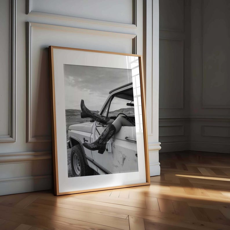 Pu&ograve; includere: Fotografia in bianco e nero incorniciata di stivali da cowboy che sporgono dal finestrino di un camion bianco. Gli stivali hanno cuciture decorative. Lo sfondo mostra un paesaggio sotto un cielo nuvoloso.