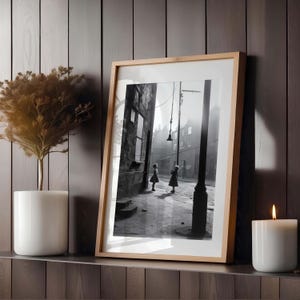 May include: Black and white photograph of two children playing in a narrow street. The photo is framed in a light wood frame and displayed on a dark wood surface.