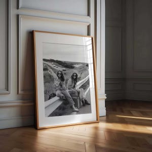 May include: Black and white framed print featuring two people sitting on a highway guardrail, one holding a sign that reads "TAKE ME ANYWHERE." They are wearing sunglasses and casual clothing. The print is in a wooden frame.