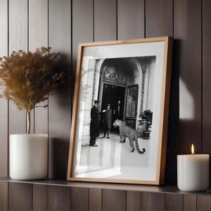 Op de afbeelding: Zwart-witfoto van twee mannen in formele kleding die voor een grote deur staan met een luipaard dat naar hen toe loopt. De foto is ingelijst in een houten lijst.