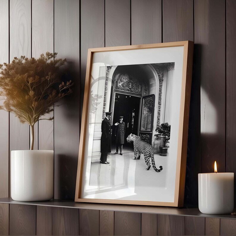 Op de afbeelding: Zwart-witfoto van twee mannen in formele kleding die voor een grote deur staan met een luipaard dat naar hen toe loopt. De foto is ingelijst in een houten lijst.