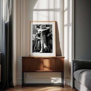 May include: A framed black and white photograph of a cowboy on a horse, wearing a hat and boots. The photograph is displayed on a wooden cabinet with a black metal frame, set against a neutral-toned wall.