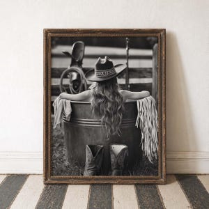May include: Monochrome framed print featuring a woman in a cowboy hat, relaxing in a metal tub. A pair of cowboy boots are placed in front of the tub. The background shows a fence and a saddle.