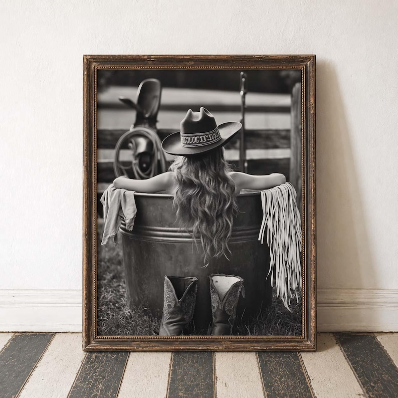 May include: Monochrome framed print featuring a woman in a cowboy hat, relaxing in a metal tub. A pair of cowboy boots are placed in front of the tub. The background shows a fence and a saddle.