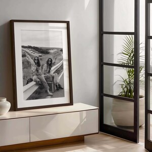 May include: Black and white framed print featuring two people sitting on a guardrail with a sign that reads "TAKE ME ANYWHERE". The print is displayed on a white cabinet with a dark brown frame.