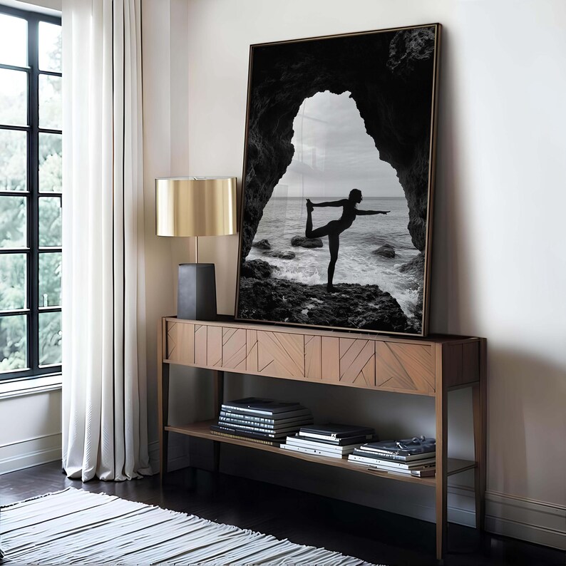 K&ouml;nnte beinhalten: Gerahmtes Schwarz-Wei&szlig;-Foto einer Person, die Yoga in einem nat&uuml;rlichen Torbogen mit Blick auf das Meer macht. Das Foto wird auf einem Holztisch mit einer goldenen Lampe ausgestellt.