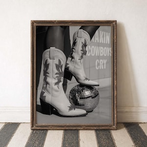 Op de afbeelding: Zwart-wit ingelijste print met lichtgekleurde cowboylaarzen met decoratieve stiksels, rustend op een discobal. De achtergrond bevat de tekst "MAKIN COWBOYS CRY".