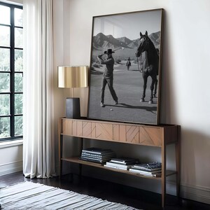 May include: A framed black and white photograph of a golfer swinging a club on a golf course, with a horse and rider in the background. The photograph is displayed on a wooden console table with a gold lamp.