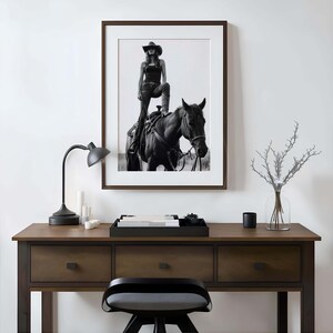 May include: Black and white framed photograph of a person in a cowboy hat standing on a horse. The photograph is hanging above a wooden desk with a black lamp, candles, and a tray.