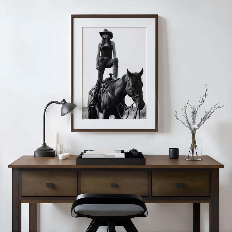 May include: Black and white framed photograph of a person in a cowboy hat standing on a horse. The photograph is hanging above a wooden desk with a black lamp, candles, and a tray.