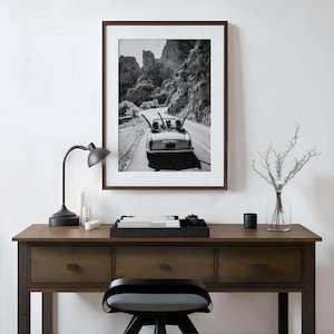 Puede incluir: Impresi&oacute;n en blanco y negro enmarcada de un coche descapotable vintage conduciendo por una carretera sinuosa a trav&eacute;s de un paisaje monta&ntilde;oso. La impresi&oacute;n se muestra sobre un escritorio de madera con una l&aacute;mpara negra, velas y una bandeja decorativa.