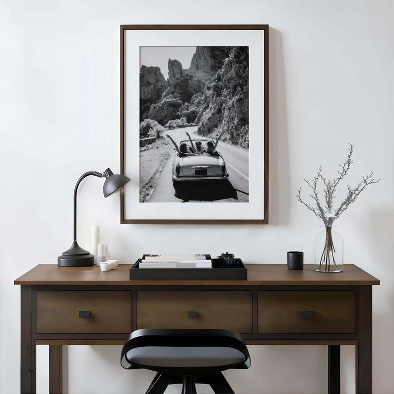 Puede incluir: Impresi&oacute;n en blanco y negro enmarcada de un coche descapotable vintage conduciendo por una carretera sinuosa a trav&eacute;s de un paisaje monta&ntilde;oso. La impresi&oacute;n se muestra sobre un escritorio de madera con una l&aacute;mpara negra, velas y una bandeja decorativa.