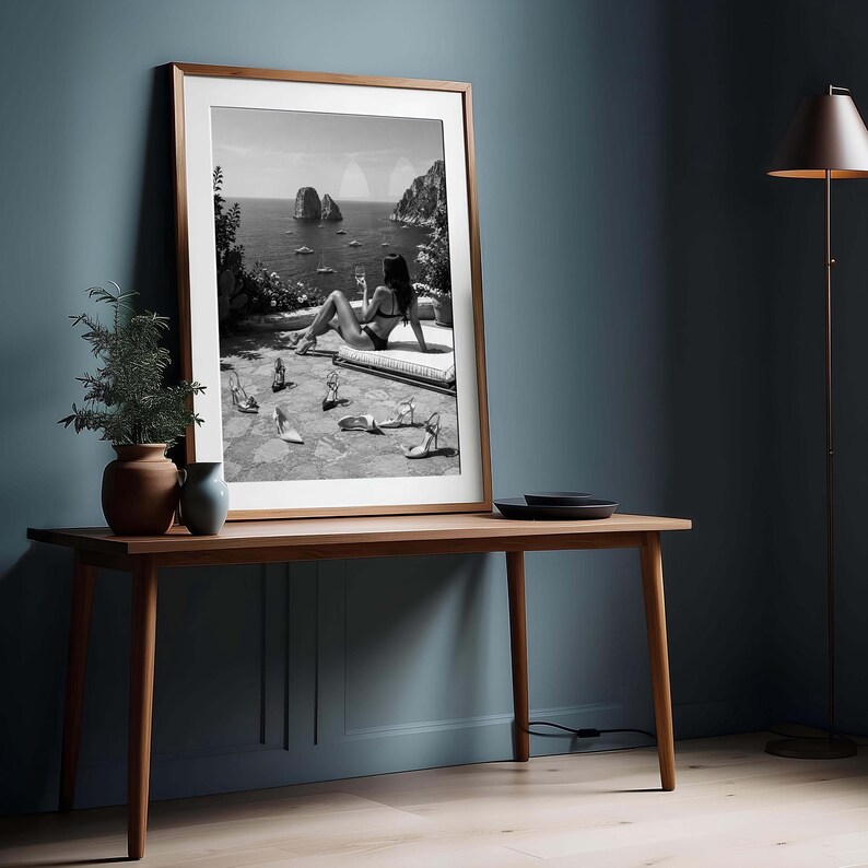 May include: A framed black and white photograph depicting a coastal scene with a figure lounging on a terrace overlooking the sea, surrounded by several pairs of high-heeled shoes scattered on the ground, presented in a wooden frame.