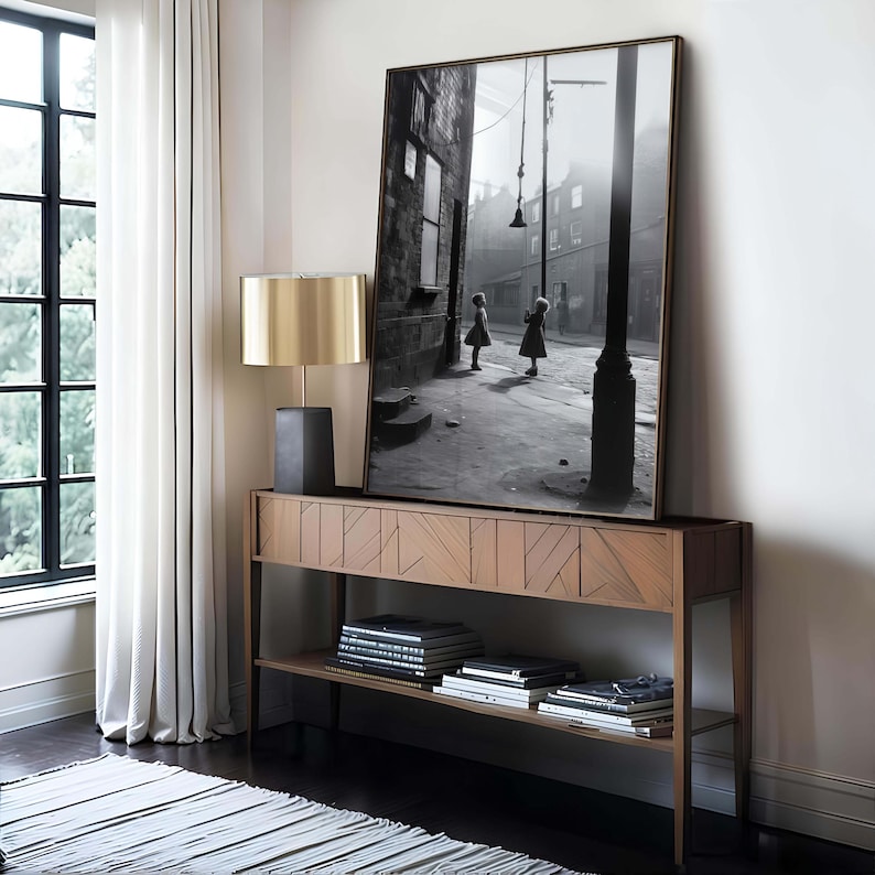 May include: A black and white photograph of two girls standing in a street. The photo is framed and hanging on a wall above a wooden console table with books on the shelf.