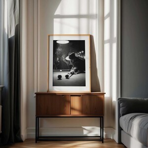 May include: A framed monochrome print of a person playing snooker, displayed on a wooden cabinet. The print shows a person in a hat, smoking, and taking a shot. The cabinet has two doors and a black metal frame.