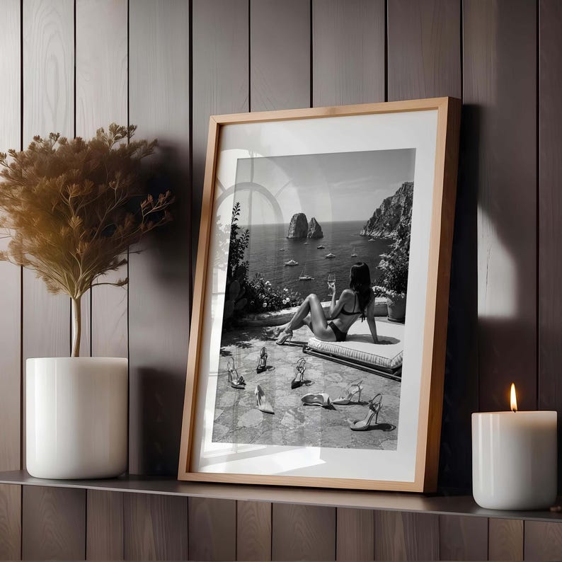 May include: A framed black and white wall art print featuring a person in a bikini sitting on a terrace overlooking the sea with rock formations and boats. Several high-heeled shoes are scattered on the stone floor in the foreground.