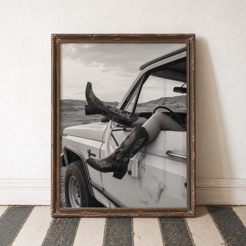 Pu&ograve; includere: Fotografia in bianco e nero incorniciata di stivali da cowboy che sporgono dal finestrino di un camion bianco. Gli stivali hanno cuciture decorative. Lo sfondo mostra un paesaggio rurale.