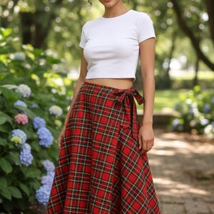 Può includere: Una donna indossa un top bianco e una gonna a quadri rossi. La gonna è a portafoglio con un fiocco in vita. Il motivo a quadri include linee rosse, nere e verdi. Lo sfondo è un giardino con alberi e fiori.
