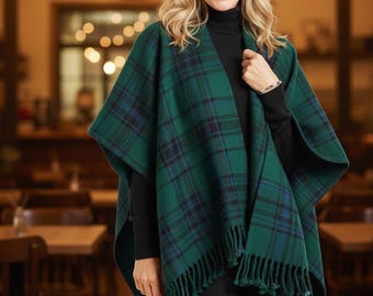 May include: A green and blue plaid cape with fringe trim. The cape is draped over a black turtleneck and black pants. The background is a blurred interior with warm lighting.