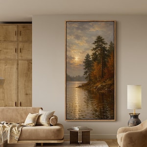 May include: A vertical framed landscape painting depicting a sunset over a calm lake with pine trees and autumn foliage on the shore. The artwork is displayed on a beige wall above a tan sofa, a wooden coffee table, and a beige armchair.