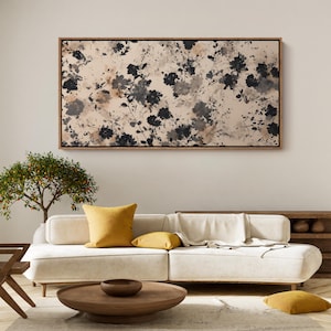 May include: A large rectangular framed canvas art piece with a floral pattern in black, gray, and beige tones. The artwork hangs above a white sofa with yellow pillows. A wooden coffee table and a small tree are in the foreground.