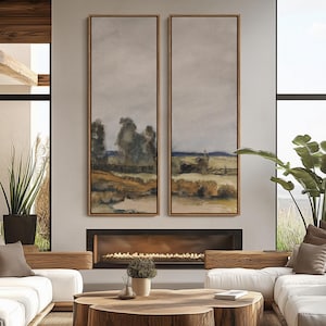 May include: Two vertical landscape paintings with wooden frames. The artwork depicts a rural scene with trees, fields, and a cloudy sky. The paintings are displayed above a fireplace in a modern living room setting.