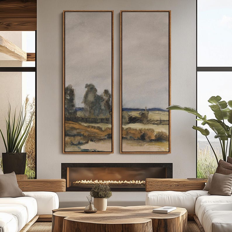 May include: Two vertical landscape paintings with wooden frames. The artwork depicts a rural scene with trees, fields, and a cloudy sky. The paintings are displayed above a fireplace in a modern living room setting.