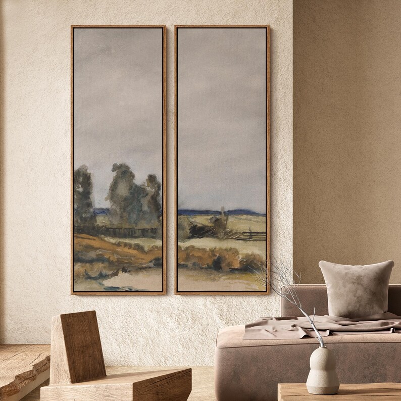 May include: Two framed landscape paintings with a muted color palette. The artwork depicts a field with a fence, trees, and a cloudy sky. The frames are a warm brown color, complementing the neutral tones of the artwork.