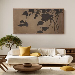 May include: A framed canvas art piece featuring a dark silhouette of a plant with large leaves and vines against a tan background. The artwork is displayed above a white sofa with yellow pillows, a wooden coffee table, and a small tree.