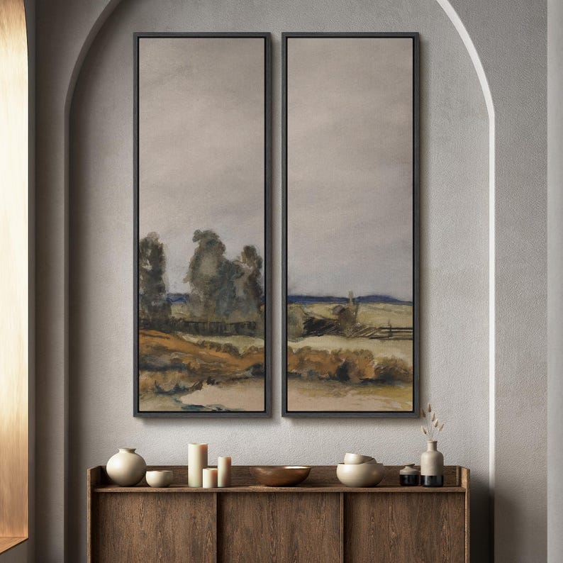 May include: Two framed landscape paintings with a muted color palette of gray, brown, and blue. The artwork depicts a rural scene with trees, fields, and a fence. The paintings are displayed above a wooden cabinet with decorative items.