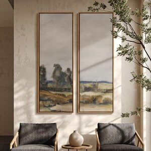 May include: Two framed landscape paintings. Each painting features a muted color palette with a field, trees, and a cloudy sky. The frames are a light wood color. The paintings are displayed above two gray armchairs.
