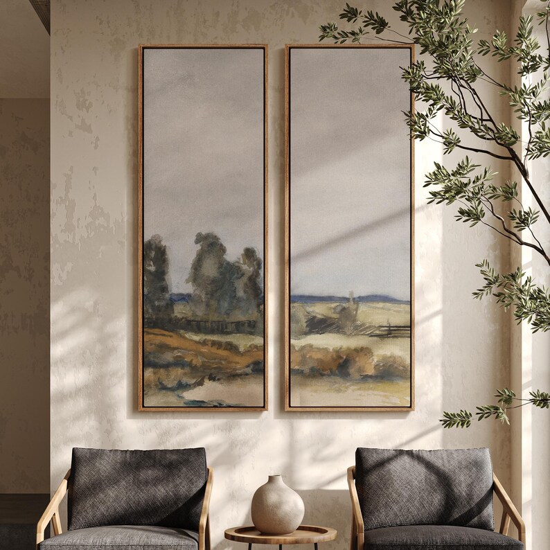 May include: Two framed landscape paintings. Each painting features a muted color palette with a field, trees, and a cloudy sky. The frames are a light wood color. The paintings are displayed above two gray armchairs.