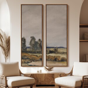 May include: Two framed landscape paintings. Each features a muted color palette with a field, trees, and a cloudy sky. The frames are a warm brown color. The artwork is displayed above a wooden cabinet.