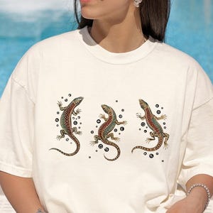 Könnte beinhalten: Ein cremefarbenes T-Shirt mit kurzen Ärmeln und einem Grafikdruck von drei Eidechsen in Grün-, Braun- und Rottönen, umgeben von kleinen schwarzen Punkten. Das T-Shirt hat einen Rundhalsausschnitt und eine lockere Passform mit Reptilienmotiv.