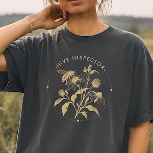 Könnte beinhalten: Ein anthrazitfarbenes Kurzarm-T-Shirt mit einer goldfarbenen kreisförmigen Grafik auf der Brust. Das Design zeigt eine Biene auf einer Blume, umgeben von botanischen Illustrationen und dem Text HIVE INSPECTOR in einem kreisförmigen Rahmen.