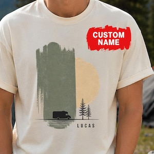 May include: A cream-colored short-sleeve t-shirt featuring a graphic print of a camper van, two pine trees, and a yellow sun against a sage green brushstroke background. The text CUSTOM NAME appears in red, and the name LUCAS is printed in black.