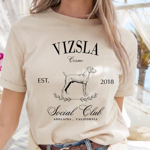Op de afbeelding: Beige t-shirt met de tekst "VIZSLA Cosmo Social Club" en een afbeelding van een hond. Het shirt bevat ook de tekst "EST. 2018" en "ADELAIDA, CALIFORNIA". Het shirt is gemaakt van zacht materiaal.