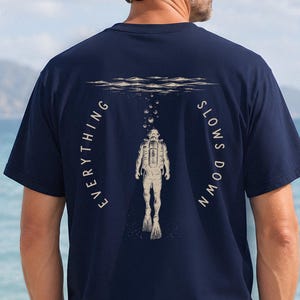 Peut inclure: Un t-shirt bleu marine à manches courtes avec un motif au dos représentant un plongeur sous-marin et des bulles remontant vers la surface. Le texte "EVERYTHING SLOWS DOWN" est imprimé en arc de cercle autour du plongeur en couleur crème.