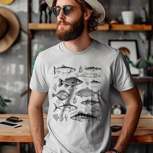 Puede incluir: Camiseta gris claro con una ilustraci&oacute;n detallada en blanco y negro de varias especies de peces. El dise&ntilde;o incluye diferentes tipos de peces, algas marinas y peque&ntilde;as plantas acu&aacute;ticas. Una prenda casual para el d&iacute;a a d&iacute;a.